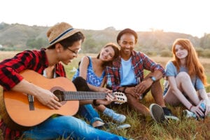 Group of happy relaxed young people drinking beer and playing guitar outdoors